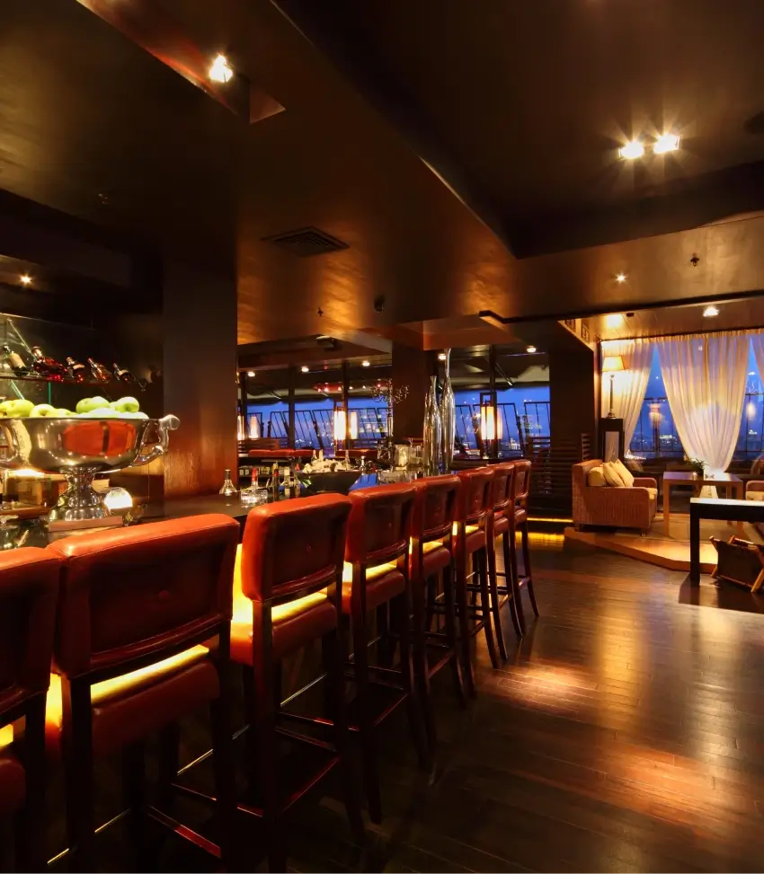An empty, elegantly lit bar area features red-cushioned stools along a polished counter, with a view into a comfortable lounge seating area.
