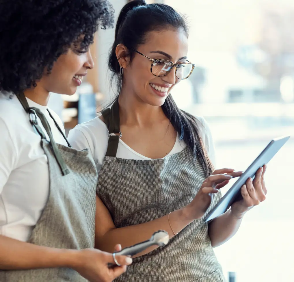 Two smiling café employees wearing aprons looking at a tablet together while working.