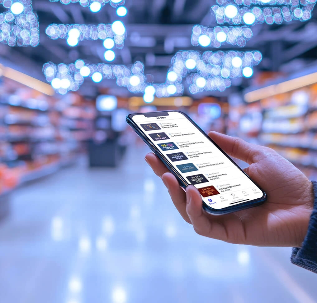 A hand holding a smartphone displaying an app in a blurred grocery store aisle with bright overhead lighting.