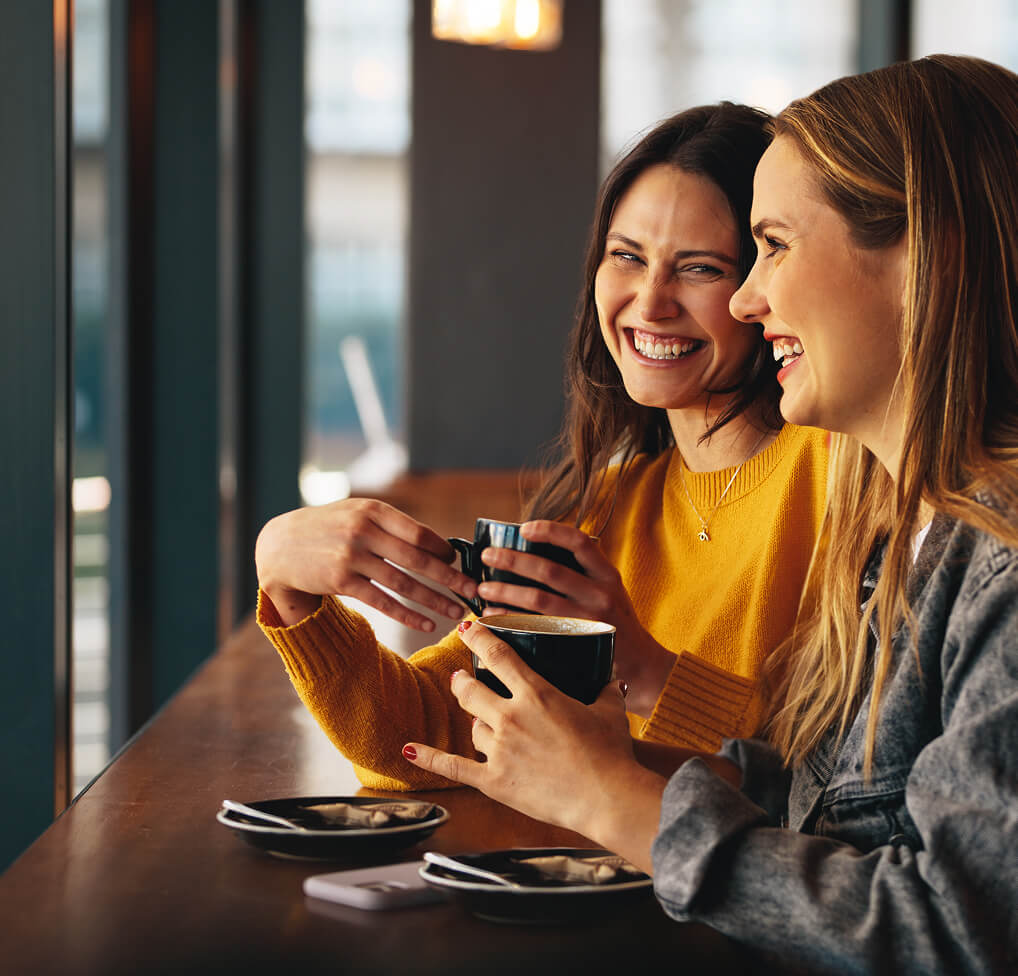 Two friends laughing and drinking coffee in a cafe with SiriusXM playing.
